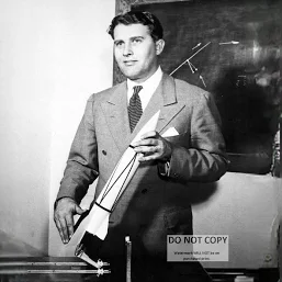Man in a suit holding a model rocket, standing in a classroom or lab with a chalkboard behind him.