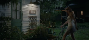 Woman in a light dress walks past a white wooden house with a lit 'Office' sign and a bicycle in a garden at dusk.