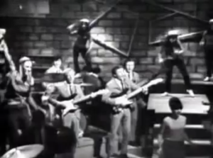 Black-and-white photo of a mid-20th-century band performing on a brick-walled stage, with guitarists and dancers on risers behind them.