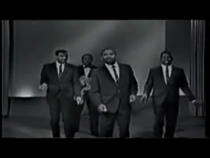 Five men in dark suits walk toward the camera on a dimly lit stage in a black-and-white video.