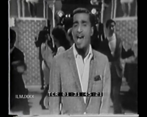 Black-and-white clip of a man in a light suit singing on stage with backup dancers behind him.