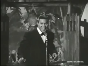 Male singer in a tuxedo performing on a vintage TV stage with a microphone in front of a jungle-set backdrop.
