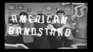 Black-and-white still of teenagers dancing in a school hallway with lockers, overlaid by large letters spelling 'American Bandstand.'