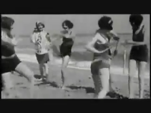 Black-and-white photo of a group of women dancing on a sunny beach in vintage swimsuits on the sand by the sea.