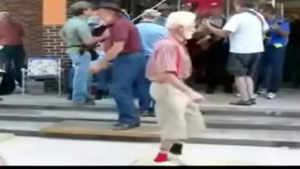 Elderly man with white hair dancing on a sidewalk near a crowd outside a store.