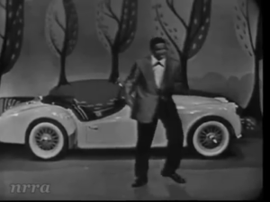 Man in a suit dancing beside a white vintage convertible on a stage with stylized trees in a black-and-white performance setting (nrra watermark). عدة alternative text.