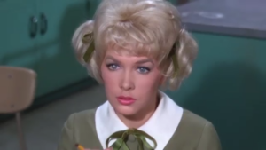 Portrait of a blonde woman in a vintage olive-green dress with a white collar and green bow, looking toward the camera in a retro setting.