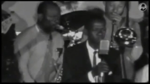 Black-and-white photo of a man in a suit singing into a vintage microphone on stage, with band members and a saxophonist behind him.