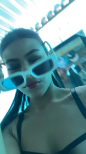 Close-up selfie of a woman wearing oversized white-framed blue sunglasses under blue neon lighting, with a tattoo on her shoulder visible.