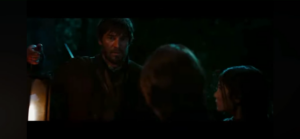 Bearded man in a dark outdoor scene points a knife toward a person off-screen while two others watch.