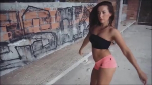 Woman in a black tube top and pink shorts walks along a graffiti-covered wall outdoors, turning toward the camera.