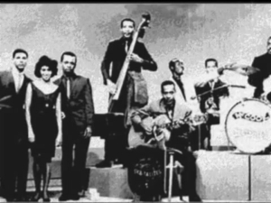 Vintage black-and-white photo of a jazz band on stage with bass, guitar, drums and other musicians posing together on a raised platform.