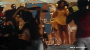People dancing in a crowded outdoor party, colorful clothing and movement visible across the scene.