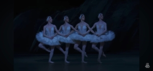 Four ballerinas in white tutus hold hands in a synchronized line on a blue-lit stage, with a Royal Ballet & Opera logo in the corner.