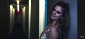 Woman in a sparkling sequined dress leans against a blue wall in a dim hallway, smiling at the camera; Vevo logo in the corner.