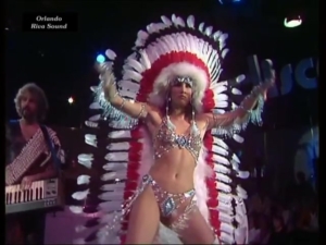 Female dancer in a sparkling bikini and large red-and-white feather headdress performing on a stage, with a keyboardist visible in the background.