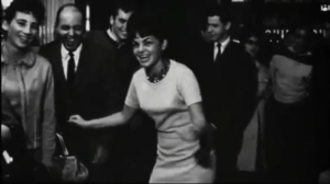 Black-and-white photo of a woman in a light dress dancing joyfully among onlookers in formal attire.