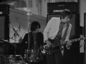 Black-and-white photo of a live band on stage: a guitarist in a light jacket and scarf playing an electric guitar while a drummer sits at a drum kit nearby.