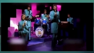 A vintage-style band in matching blue suits performs on a colorful stage, with a drummer behind a white‑drum kit in back and singers at microphones in front.