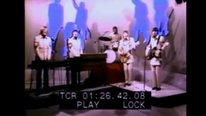 Band performance on a stage: keyboard/piano in front, drummer in the back, two guitarists or singers on sides, with a blue-pink abstract backdrop.