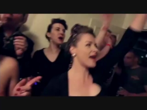 People at a lively party, cheering with hands raised and singing along in a crowded room.