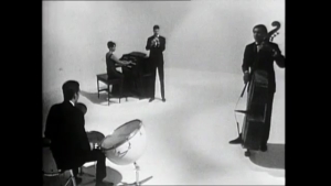 Black-and-white studio scene with five musicians: pianist at piano, drummer at kit, trumpeter singing into a mic, vocalist, and a standing double bassist.