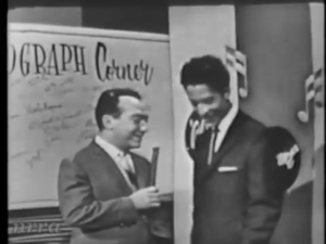 Two men in suits on a stage; one hands a rolled document or diploma to the other, with a banner behind them that partially reads 'Center' (likely a venue banner).