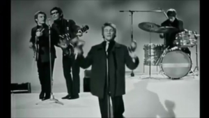 Black-and-white band performance with a lead singer at the mic center stage, guitarists to the left, and a drummer on the right.