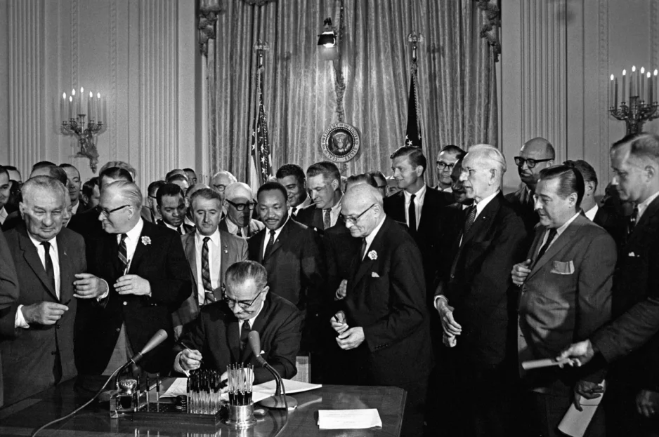 A man signs documents at a desk as a crowd of suited men surrounds him; presidential seal and flags behind.