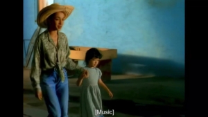 Woman wearing a wide straw hat and patterned blouse walks hand-in-hand with a young girl along a blue seascape background.