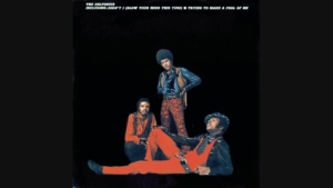 Album cover style image: three men in 1970s outfits posed against a black background, with an abstract orange shape at the bottom and white text at top.