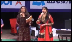 Two women in traditional outfits speak on a stage with microphones, notes in hand, banners and chairs in the background.