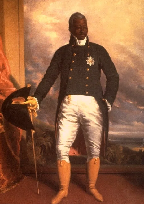 Wax figure of a dark-skinned naval officer in a black coat with brass buttons, white breeches, and tall boots, holding a hat and cane.