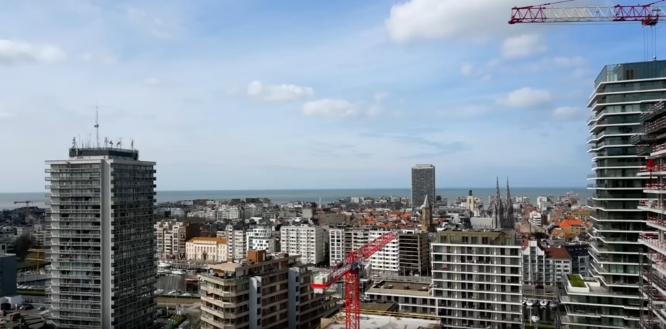 City skyline with tall modern buildings, red construction crane, and the sea on the horizon under a blue sky with scattered clouds